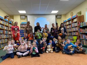 Duża grupa dzieci i Panie w bibliotece. Dzieci trzymają w rękach pluszowe zabawki.