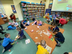 Duża grupa dzieci siedzi na dywanie w bibliotece. Niektóre oglądają książki, inne grają w gry.
