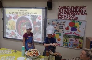 Dziewczynka i chłopiec ubrane w czapki kucharskie i fartuszki, stojący za stołem. Chłopiec wyciska z sokowirówki sok. Przed dziewczynką stoi miska z owocami i warzywami, kubki, talerzyki oraz trzy łyżeczki.
