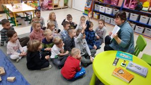 Grupa dzieci siedzi w rozsypce na dywanie. Przed dziećmi na zielonym krzesełku siedzi poetka i pokazuje książkę. Obok na zielonym okrągłym stoliku położone są trzy książki, zakładki oraz pieczątka.
