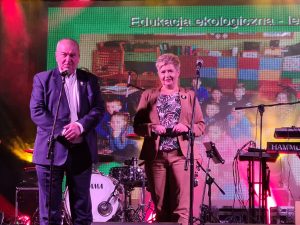 Na scenie stoi kobieta i mężczyzna. Za nimi widoczne są instrumenty muzyczne.