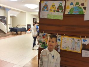 Chłopiec w białej koszuli, za nią powieszone prace konkursowe.