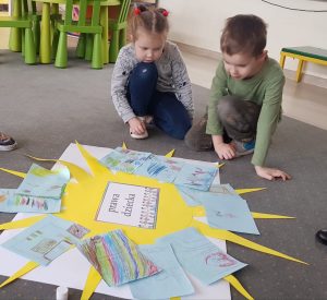 Dziewczynka i chłopiec siedzą na szarym dywanie w przedszkolnej sali. Przed chłopcami leży arkusz białego papieru, a na nim naklejone żółte słońce. Na jego promieniach są kartki z rysunkami.