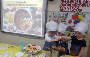 Trzy dziewczynki ubrane w fartuszki i czapki kucharskie, wyciskajace sok z sokowirówki. Na stole stoi miska z owocami, kuki oraz trzy łyżeczki.