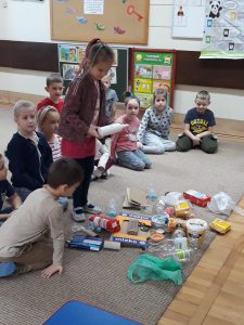 Dzieci siedzą na dywanie , przed nimi leżą rozrzucone śmieci, które dzieci muszą posegregować i wrzucić do odpowiedniego worka.