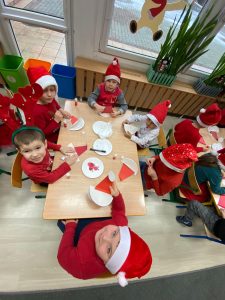 Dzieci ubrane na czerwono z czapeczkach mikołaja siedzą przy stoliku i wykonują prace plastyczną.
