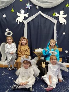 5 dzieci siedzi na dywanie lub krześle. Na środku stoi żłobek po bokach dziewczynka w przebraniu Maryi i chłopiec w przebraniu Józefa. Przed nimi siedzą dziewczynki przebrane za aniołki.