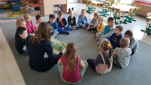 Grupa dzieci i Pani siedzą na dywanie w przedszkolnej sali i słuchają czytanego opowiadania.