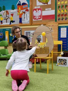 Dziewczynka rzucza piłeczką w słoik z kartonu z napisem MIÓD i wyciętą w środku dziurą.