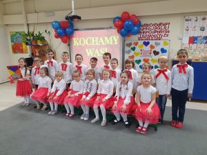 Grupa dzieci ustawiona w dwóch rzędach. W pierwszym rzędzie dziewczynki siedzą na krzesełkach. W tle na tablicy interaktywnej napis "Kochamy Was.".