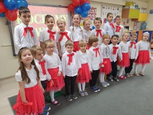 Grupa dzieci ustawiona w dwóch rzędach. W tle na tablicy interaktywnej napis "Kochamy Was.".
