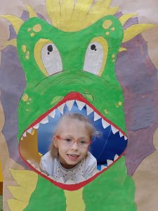 Dziewczynka siedzi na plakatem przedstawiającym dinozaura z otwartą buzią.