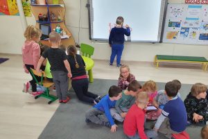 chłopiec w granatowej bluzie rysuje na tablicy interaktywnej. Pięcioro dzieci znajduje się przy zielonym stoliku, na którym jest laptop. Pozostałe dzieci siedzą na dywanie z tabletem.