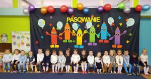 Duża grupa dzieci ubranych na galowo siedzi na ławce w rzędzie.