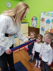 Pani Dyrektor przykłada dziecku do ramienia dużą, drewnianą kredkę.