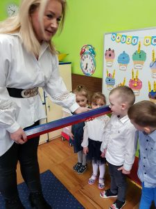 Pani Dyrektor przykłada dziecku do ramienia dużą, drewnianą kredkę.