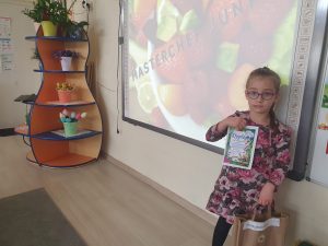 Dziewczynka ubrana w sukienkę w kolorowe kwiaty, stoi przy tablicy interaktywnej i trzyma dyplom oraz upominek w papierowej torbie.