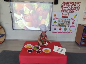 Dziewczynka ubrana w sukienkę i kolorowy fartuszek siedzi przy stoliku i układana na talerzu z pokrojonych owoców pawia. Na stole położony jest czerwony obrus oraz talerze z owocami takimi jak maliny, borówki, ananas oraz jasne i ciemne winogrono. Na głowie dziewczynka ma czapkę kucharza.