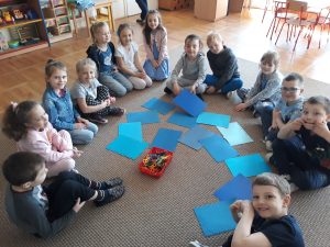 Dzieci siedzą na dywanie. Na środku leżą niebieskie kartki oraz koszyk z nożyczkami.