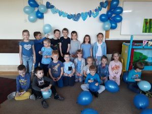 Grupa dzieci ubrana na niebiesko ustawiona w dwóch rzędach. dzieci w pierwszym rzędzie siedzą , przed nimi leżą niebieskie balony. W tle widoczna jest dekoracja z niebieskich serduszek i balonów.