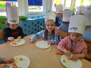 Dzieci w czapkach kucharza kroją marchewki i jabłka