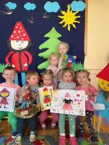Grupa dzieci ustawiona w dwóch rzędach. Dzieci trzymają wykonane przez siebie prace konkursowe. W tle za nimi dekoracja krasnal Hałabała, choinka słońce, chmury i jaskółki.