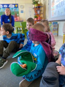 Chłopiec siedzi na dywanie z zawiązanymi oczami, na ręce ma założoną kuchenną rękawice. Chłopiec trzyma w niej marchewkę.