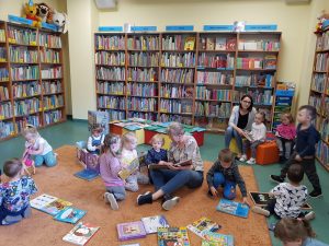 Dzieci i Panie siedzą na dywanie w bibliotece. Każde dziecko czyta książeczkę.