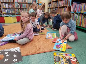 Dzieci i Panie siedzą na dywanie w bibliotece. Każde dziecko czyta książeczkę.