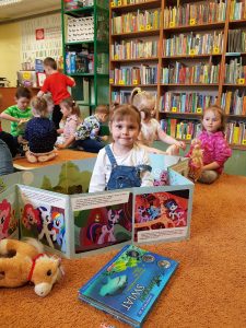 Dzieci siedzą na dywanie w bibliotece. Każde dziecko czyta książeczkę.