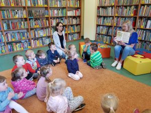 Dzieci siedzą na dywanie w bibliotece. Patrzą na Panią która czyta im książkę.