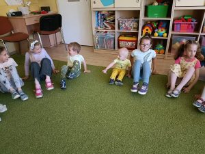 Dzieci i Pani siedzą na dywanie w kole i tunią nogami