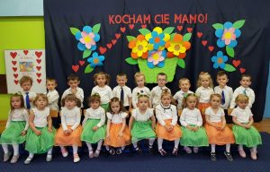 Duża grupa dzieci z odświętnych strojach . Za nimi tło z napisem kocham cię mamo i kwiatami