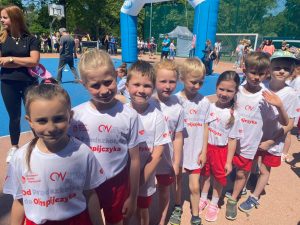 Ośmioro dzieci ustawionych w jednym rzędzie jeden za drugim na boisku, ubranych w równe stroje sportowe. W tle widoczni ludzie oraz przyrzady sportowe.
