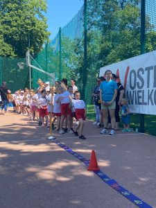 Grupa dzieci ustawionych w jednym rzędzie jeden za drugim na boisku, ubranych w równe stroje sportowe. Po prawej stronie stoi kilkoro osób dorosłych i dziecko.