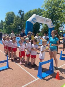 Grupa dzieci ustawionych w jednym rzędzie jeden za drugim na boisku, ubranych w równe stroje sportowe. Pierwszy chłopiec trzyma w rękach piłkę. Przed dziećmi stoi dziewczyna. W tle widoczni ludzie oraz przyrządy sportowe.