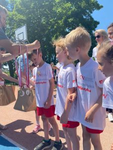 Czworo dzieci stojących obok siebie na boisku ubranych w stroje sportowe. Chłopiec po lewej stronie ma wręczany przez mężczyznę medal. Z tyłu stoją dwie panie.