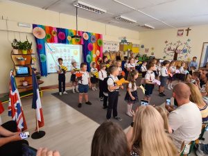 Dzieci stojące w sali w trzech rzędach. Dzieci stojące w pierwszym rzędzie trzymają motyle, w drugim biedronki, w trzecim pszczoły. Po lewej stronie stoją flagi. Przed dziećmi, na krzesełkach siedzą rodzice.