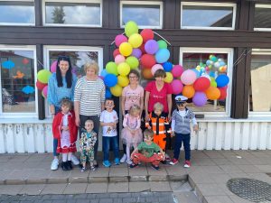 Grupa dzieci stojąca przed budynkiem przedszkola. Nad głowami wiszą kolorowe balony.