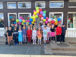 Grupa dzieci stojąca przed budynkiem przedszkola. Nad głowami wiszą kolorowe balony.