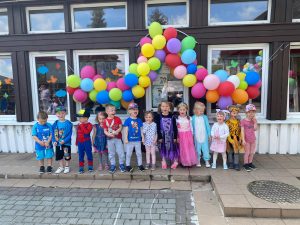 Grupa dzieci stojąca przed budynkiem przedszkola. Nad głowami wiszą kolorowe balony.