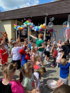 Grupa dzieci uczestnicząca w zabawach ruchowych przed budynkiem przedszkola. W tle Pani puszcza bańki mydlane. Budynek przedszkola jest udekorowany balonami.
