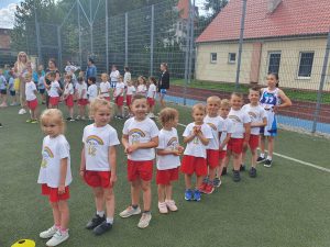 Dzieci w białych koszulkach z logo przedszkola oraz w czerwonych spodenkach, ustawieni w jednym rzędzie na boisku sportowym.