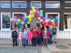 Grupa dzieci stojąca przed budynkiem przedszkola. Nad głowami wiszą kolorowe balony.