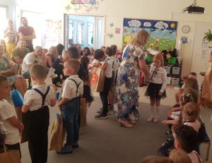 Pani Dyrektor trzymająca za rękę dziewczynkę. Po lewej stronie stoją dzieci trzymający papierowe torby z upominkami. Na ławkach siedzą rodzice.