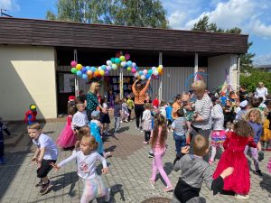 Grupa dzieci uczestnicząca w zabawach ruchowych przed budynkiem przedszkola. W tle widocznych jest 5 pań, które puszczają bańki mydlane. Budynek przedszkola jest udekorowany kolorowymi balonami.