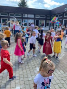 Grupa dzieci uczestnicząca w zabawach ruchowych przed budynkiem przedszkola. Budynek przedszkola jest udekorowany kolorowymi balonami.