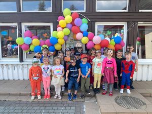 Grupa dzieci stojąca przed budynkiem przedszkola . Nad dziećmi wiszą kolorowe balony.