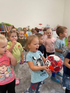 Grupa dzieci stoją na dywanie. Jedna dziewczynka trzyma piłkę.