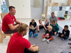 Chłopcy i nauczycielka siedzą na dywanie i słuchają 2 sportowców.